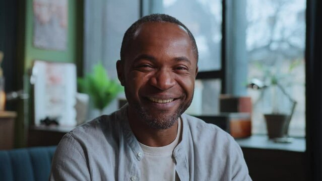 Horizontal shot of smiling handsome African American man in casual outfit looking directly at camera while sitting in front of laptop. Cozy modern interior on blurred background