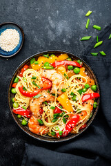 Stir fry egg noodles with shrimps, paprika, green pea, chives and sesame seeds bowl. Asian cuisine dish. Black kitchen table background, top view