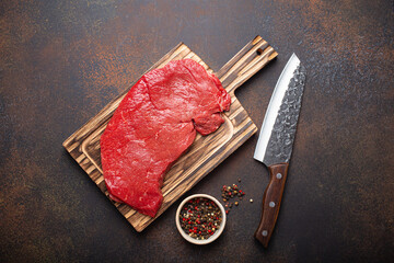 Raw uncooked top round beef steak on wooden cutting board with big kitchen knife and pepper on dark...