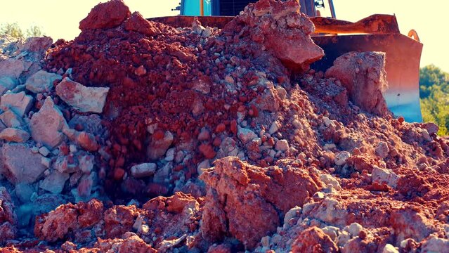 Static shot of bulldozer pushing large red and blue clay stones towards camera. Large metal blade shines and covers all frame. Stones crumles. Then dozer moves away. 50perc slow motion. 4k.