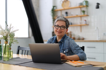 Positive woman with gray hair works on a laptop in a home interior.