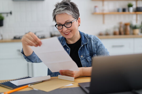 Smiling senior woman in glasses looking through mail in home interior.