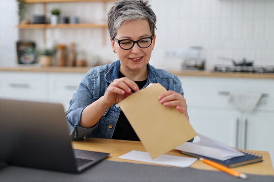 Senior woman opens a mail envelope at a workplace in a home interior. - Powered by Adobe