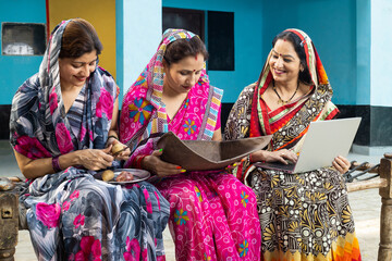 Group of traditional married indian women wearing sari do household kitchen work together at home and one lady working on laptop.