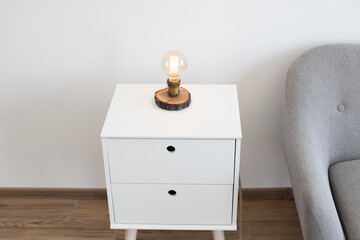 Industrial edison lamp on white contemporary bedside table near grey sofa in spacious studio room. Classic light providing dimmable warm glow in minimalistic interior of white and grey colors.