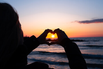 a woman arranges her hands in the shape of a heart against the background of the setting sun over the sea at sunset - obrazy, fototapety, plakaty