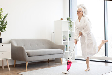 Smiling barefoot lady after shower using lightweight stick vacuum for daily clean-up of airy studio room. Joyful female homeowner saving time and energy while hoovering with easy handheld device. © sofiko14