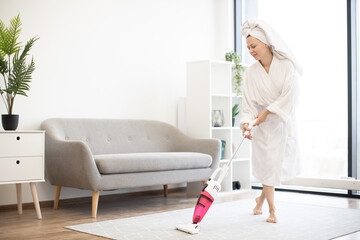 Happy woman in white robe holding upright vacuum cleaner and showing thumb-up sign while standing...