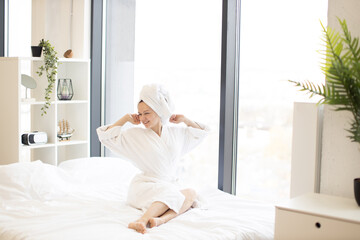Smiling female adult in white dressing gown stretching her limbs while resting on cozy bedlinen in bright room of studio apartment. Joyful housewife increasing life satisfaction by having fun at home.