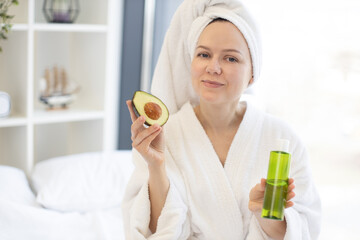 Obraz na płótnie Canvas Charming young woman in after-shower outfit enjoying exotic fruit while holding glass vial in bedroom interior. Beautiful female enhancing skin health with avocado facial oil during at-home spa day.