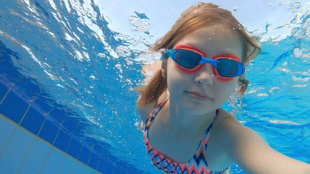 Cute little girl with goggles diving and swimming underwater in a pool. Kid taking selfie in water.