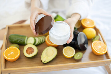 Young female opening cosmetic product with white creamy texture while sitting on soft blanket near organic beauty remedies on bed tray. Light moisturiser reducing development of skin dryness.