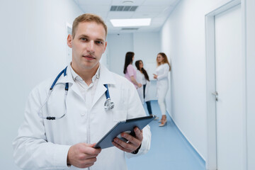 Portrait of a male doctor with a tablet in his hands inside the hospital. The Doctor looks into the camera. There is a team of doctors in the background.