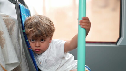 Sad child traveling by bus holding into metal bar pole. One passenger little boy sitting inside bus city transportation with upset expression