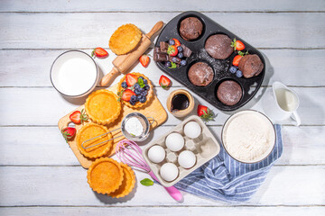 Obraz na płótnie Canvas Summer baking background with baked pastry - berry cakes, muffins, mini pie tarts, with cooking baking ingredients, flour, eggs, rolling pin, cream milk, sugar, strawberry, white wooden background