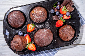 Chocolate muffins with berry. Baking muffin pan with homemade sweet dark chocolate cupcakes with cocoa sauce, sugar powder and fresh strawberry, blueberry, on white wooden background copy space