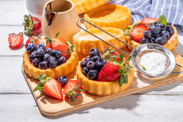 Sponge mini tart cakes with summer berry. Portioned vanilla dessert cups with fresh strawberry, blueberry, raspberry, on white wooden background copy space