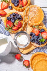 Sponge mini tart cakes with summer berry. Portioned vanilla dessert cups with fresh strawberry, blueberry, raspberry, on white wooden background copy space