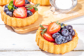 Sponge mini tart cakes with summer berry. Portioned vanilla dessert cups with fresh strawberry, blueberry, raspberry, on white wooden background copy space