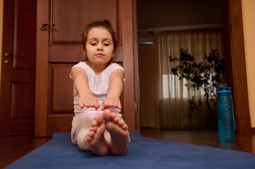 Adorable child girl doing stretching exercises, sitting barefoot on a blue gym mat while exercising...