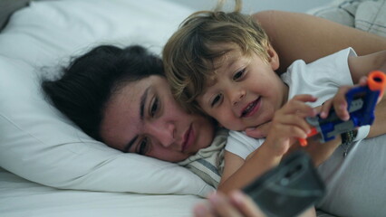 Mother and child laying in bed together looking at smartphone device. Mom embracing little boy looking at phone screen with parent. Two people