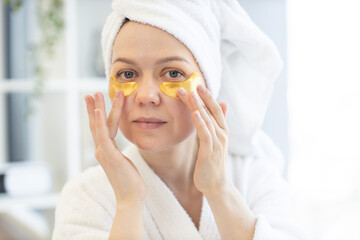 Close up view of lovely caucasian female in towel and dressing gown applying pair of eye patches after cleansing procedures in morning routine. Pretty woman taking care of delicate skin around eyes.