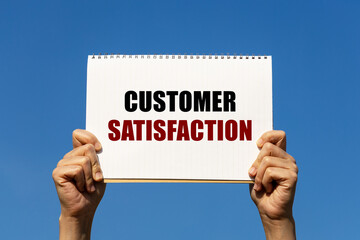 Customer satisfaction text on notebook paper held by 2 hands with isolated blue sky background. This message can be used as business concept about customer satisfaction.