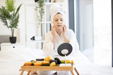 Lovely caucasian lady with towel on head fixing facial sheet mask while sitting in front of tray...