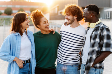 Happy multiracial friends in the city at sunset - Four young students hugging each other outdoors 