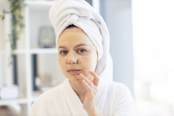 Pretty caucasian female in white robe and towel touching face with applied paper sheet mask while spending free time at home. Young person benefiting beauty care procedures in bedroom interior.