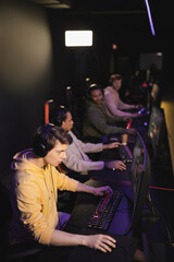 Gamer in headphones playing video game on computer near blurred interracial team in gaming club.