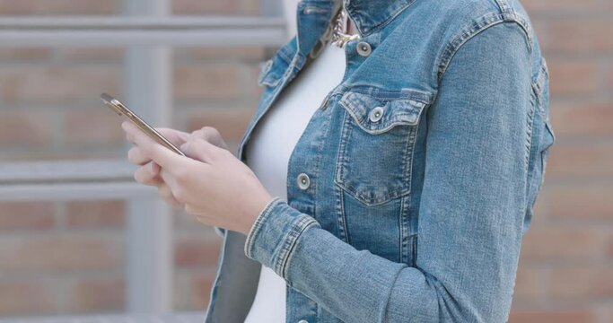 Young caucasian woman in fashionable attire and a blue jeans jacket uses smartphone to find something. No face. side view. High quality 4k footage