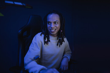 Smiling multiracial gamer with dreadlocks sitting in cyber club with lighting.