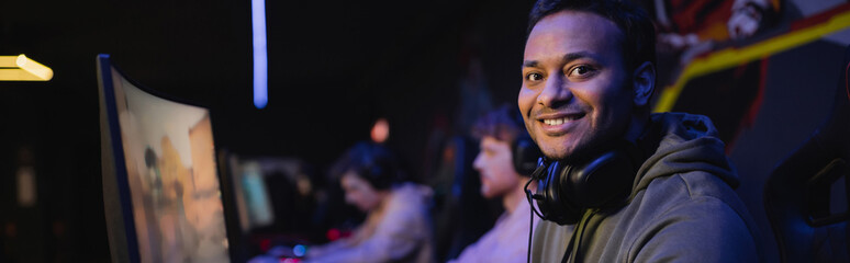 Positive indian gamer with headphones looking at camera in cyber club, banner.