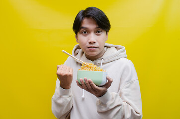 Closeup an Asian man in a beige hoodie eats instant noodles using chopsticks and a bowl, isolated over a yellow background. 