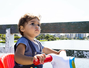 Baby sitting on a blue and red scooter on a bridge over a lake in a quaint urban location with animals, trees and buildings. Curiosity and new learning outdoors and neurological development.