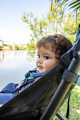 Baby sitting in a stroller in front of a lake in an urban area with trees and nature around looking towards the shore concentrated on new discoveries. Curiosity and learning new outdoors.