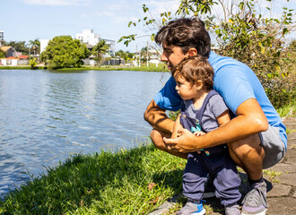 Baby beside his father in front of a lake in an urban area with fish, animals, trees and buildings in the open air. Curiosity and new outdoor learning and neurological development.
