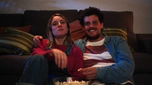 Front view of young caucasian couple smiling and having fun watching tv at late night sitting on the sofa at home, eating popcorn, turning on the television with the remote control. Love relationship