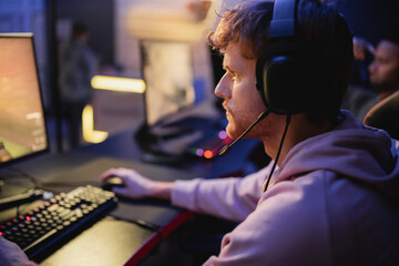 Side view of focused gamer in headphones playing video game on computer in gaming club.