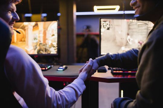 Positive gamers shaking hands near blurred computers in gaming club.