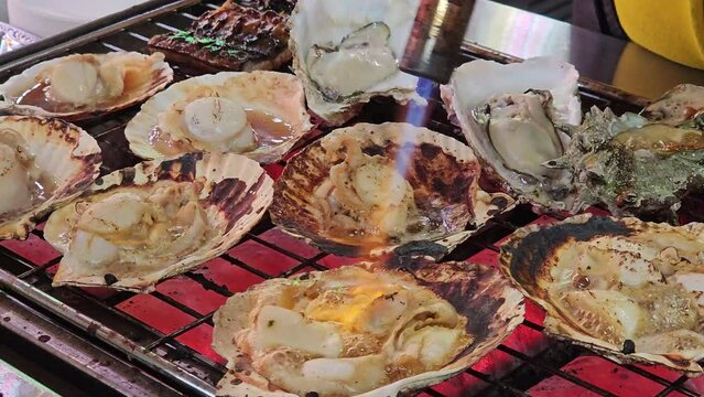 Japanese chef using hand-held burner burning to burn hotate scallops over a charcoal fire.