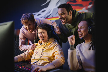 Excited interracial men looking at friend playing video game on computer in gaming club.