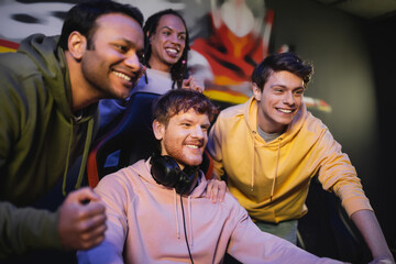 Carefree interracial team looking away near friend with headphones in gaming club.