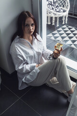 A young girl sits on the floor by the window and drinks wine.