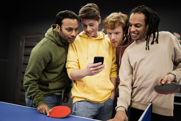 Cheerful multiethnic friends using cellphone near table tennis in gaming club.