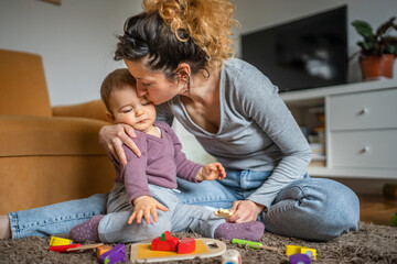 Naklejka premium One young woman sitting at home on the floor playing and kissing her one year old baby, love, family, early child development concept