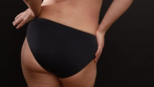 Back view of unrecognizable plus-size woman wearing black sports panties, dancing on black background, holding hands on waist, clapping, moving shaking body hips. Body positive, weight loss, dance.