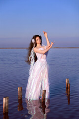 Young long haired woman in white fairy dress standing in blue water. Fantasy portrait of maiden with closed eyes on water and sky background