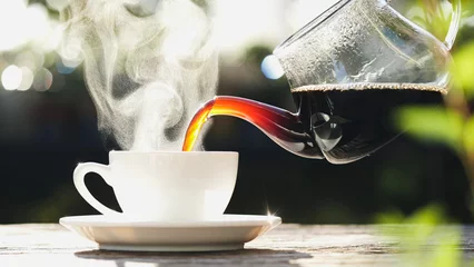 Selbstklebende Fototapeten Cafe Pouring hot black coffee to white coffee cup, mug with steaming smoke of coffee on old wooden table in morning nature outdoors, garden background. Hot Drink, Beverage Concept  © iHumnoi